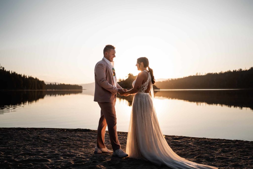 Beautiful wedding photo of Bride and Groom Staring at each other holding hands on lakeside with sunset