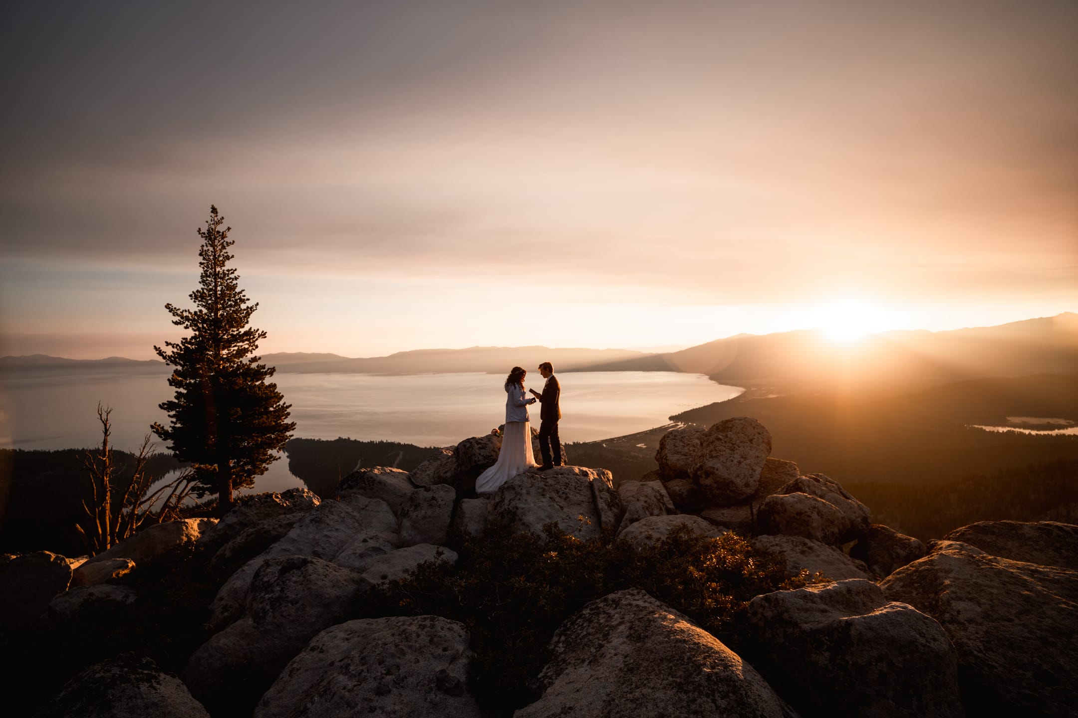 Beautiful wedding photo of Bride and Groom standing on mountain rocks with lake side view and sunset