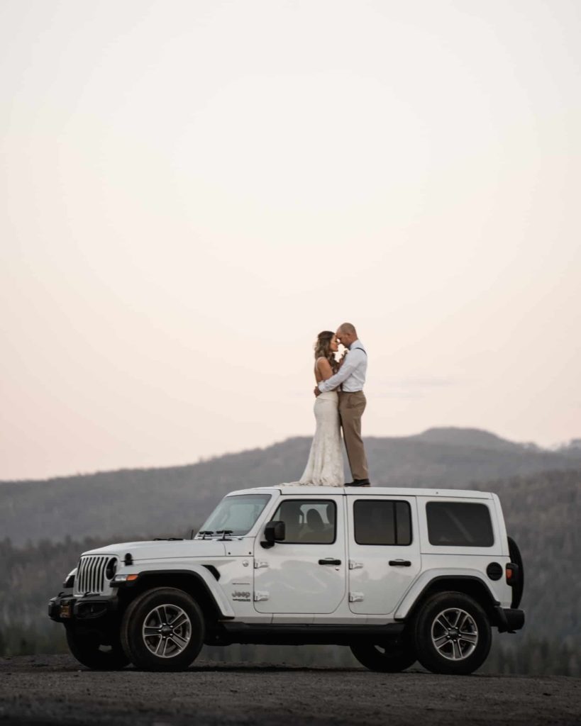 Epic elopement photo of Bride and Groom standing on a white Jeep Wrangler Hugging with mountain view