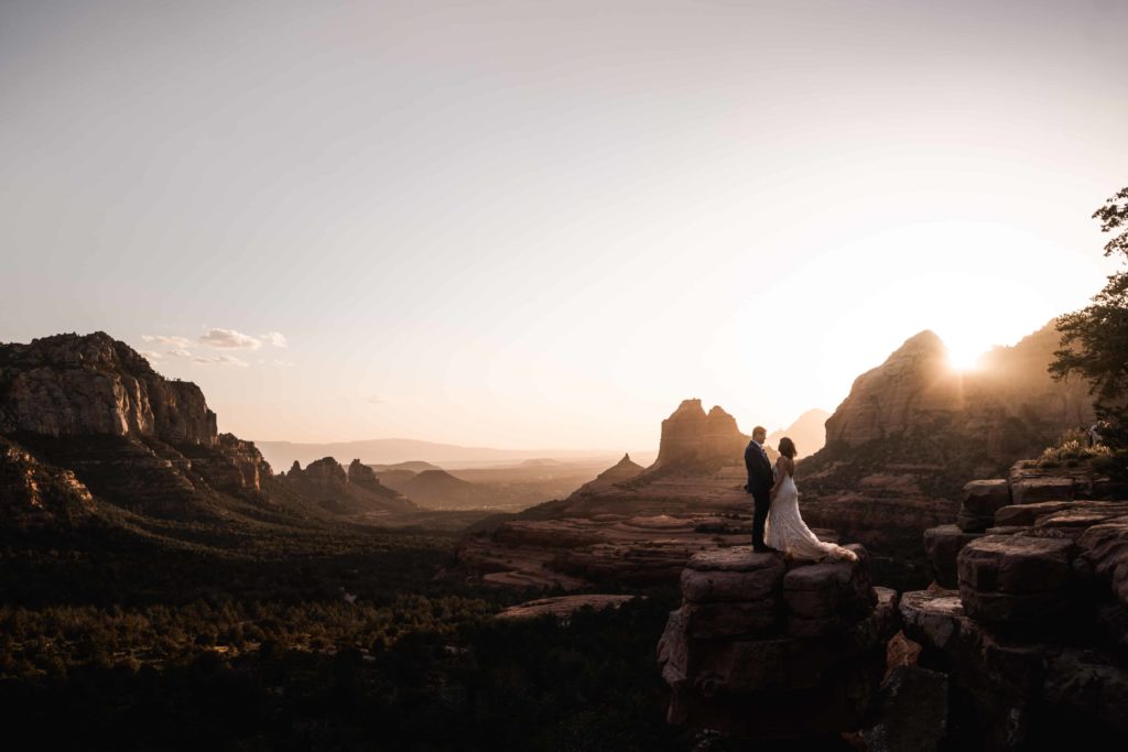 A couple eloping in Arizona, standing on the edge of a majestic red rock formation in Sedona at sunset, with panoramic views of the valley and towering buttes in the background, symbolizing adventure and intimacy in a serene desert landscape.