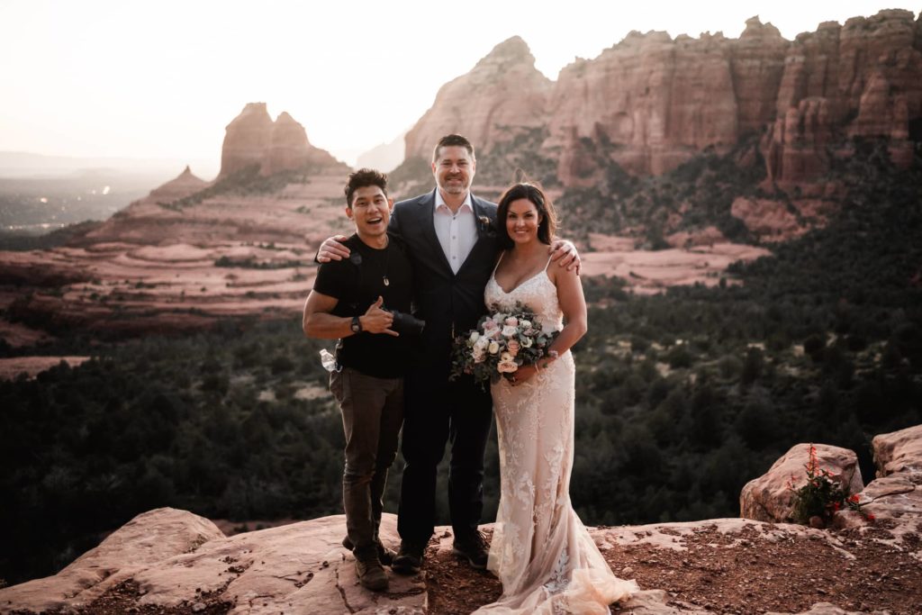 Bride and groom posing with their photographer on a Sedona cliff at sunset, celebrating their Arizona elopement with scenic red rock views in the background