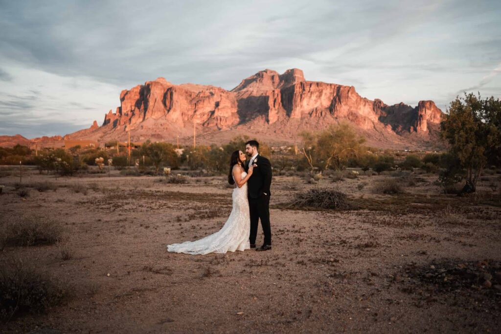 Bride and groom sharing a kiss in the vast Arizona desert, with the iconic red sandstone cliffs of the Superstition Mountains in the background, embodying a romantic elopement in the rugged landscape of Arizona, captured during the golden hour for a warm, natural ambiance.