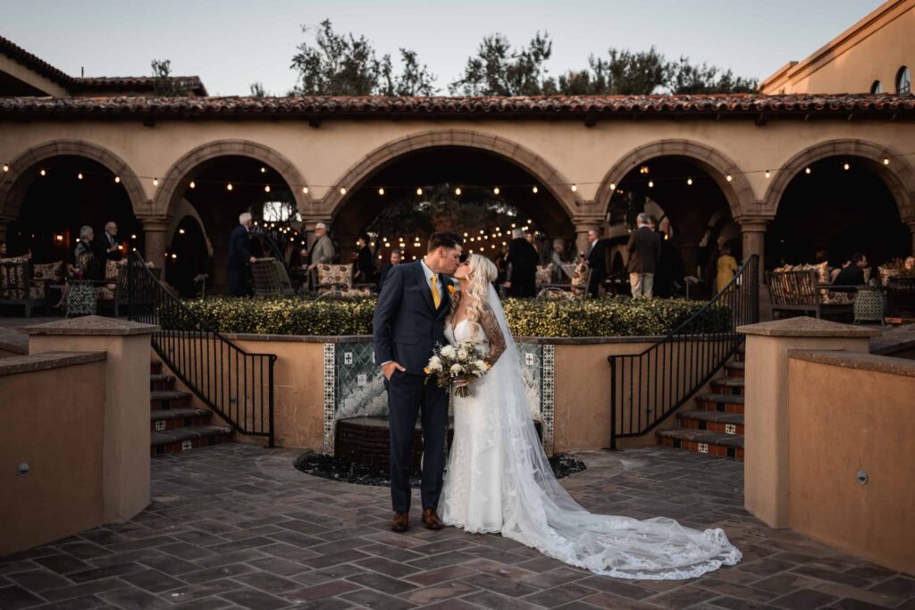 Bride and groom sharing a kiss in a charming courtyard with Spanish architecture, as guests mingle in the background under warm glowing lights. The bride's elegant lace train cascades down the steps, complementing the romantic evening atmosphere at this destination wedding