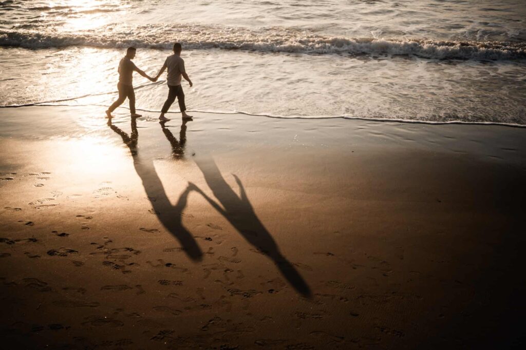 Two people holding hands and walking along the beach at sunset, with their elongated shadows reflecting on the wet sand, while gentle waves crash nearby. engagement photo