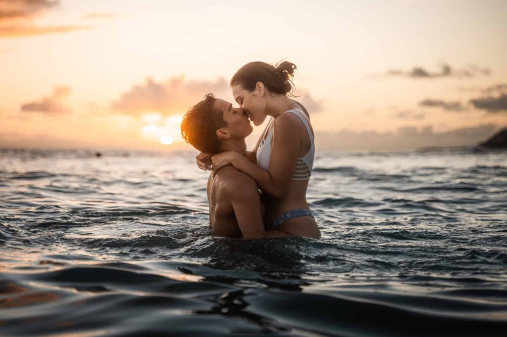 "Beach engagement photos of a couple embracing in the water during sunset. The sun casts a warm glow, illuminating their profiles. The man, with wet hair, lifts the woman slightly as they are about to share a kiss. She, wearing a striped swimsuit, holds onto his shoulders. The serene ocean surrounds them, with the sun setting in the distance, casting reflections on the water