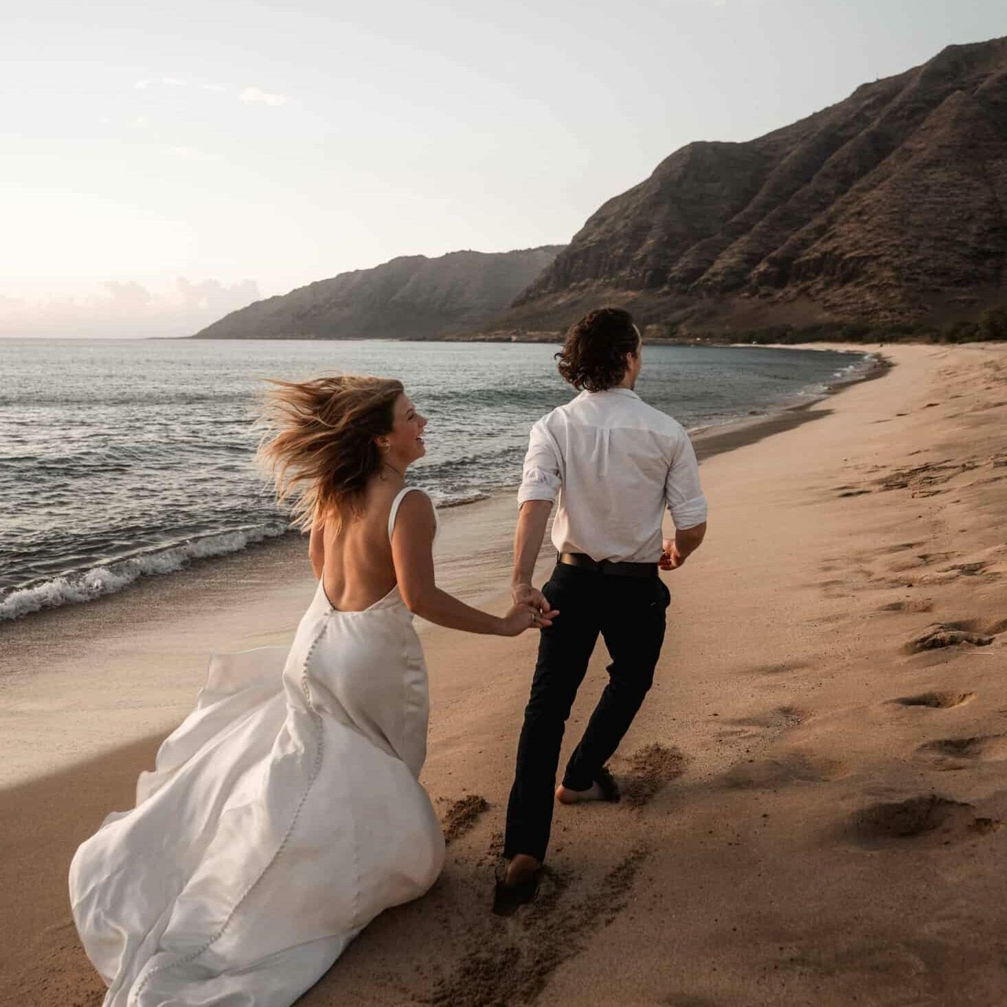 A joyful bride in a flowing white dress and a groom in a white shirt and black pants holding hands while walking on a sandy beach, with the bride's hair blowing in the wind and scenic mountains in the background