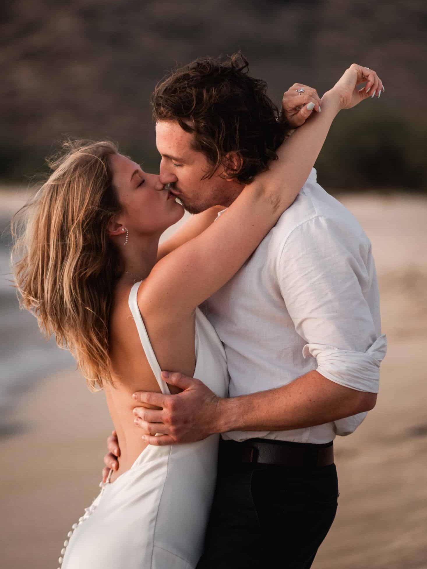 Beach engagement photos of a couple passionately embracing and kissing. The woman, wearing a white dress, has her arm draped over the man's shoulder, showing off an engagement ring. The man, dressed in a white shirt and black trousers, holds her close, with the muted colors of the beach and hills in the background