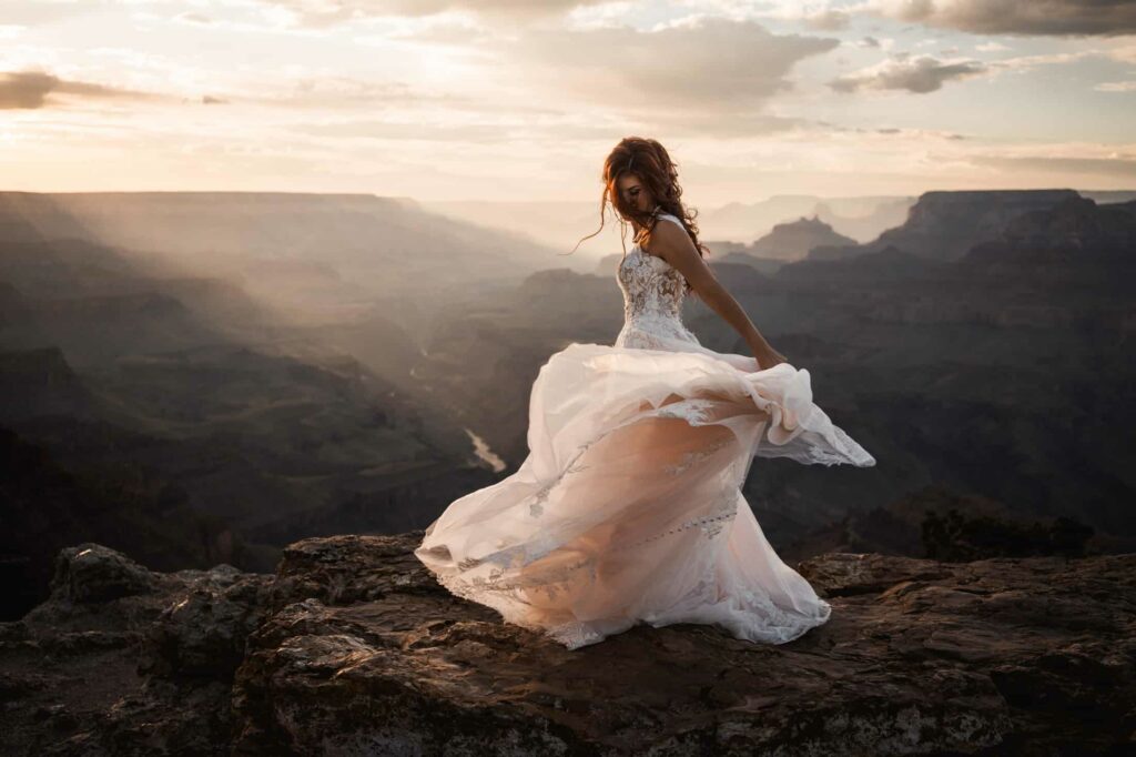 Bride in a stunning lace wedding gown standing on a rock at the Grand Canyon, with the dress flowing in the breeze and sunset illuminating the vast canyon landscape behind her, capturing romantic bridal portrait