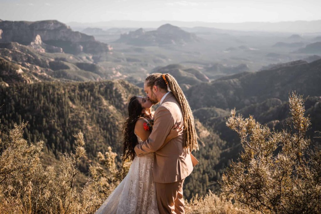 A heartfelt embrace between a bride and groom on a scenic hilltop with panoramic views of Arizona’s rugged landscape, the bride's lace dress and the groom's dreadlocks adding unique personal touches to their wedding attire, as they share a kiss amidst the natural beauty