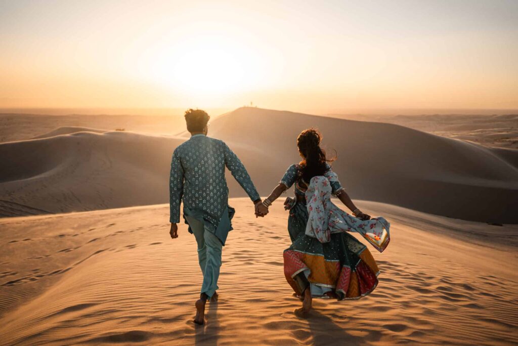 A man and woman standing on a desert dune at sunset, holding hands with their backs to the camera, silhouetted against the vibrant orange sky, creating an atmosphere of romance and adventure in the vast, serene sandscape