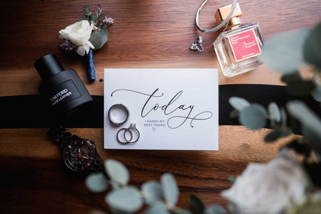A flat lay wedding setup with elegant  bride and groom's accessories, including a black wristwatch, a cologne bottle, a handwritten card stating 'Today I marry my best friend', wedding rings, a floral boutonniere, and a bottle of perfume. for their elopement
