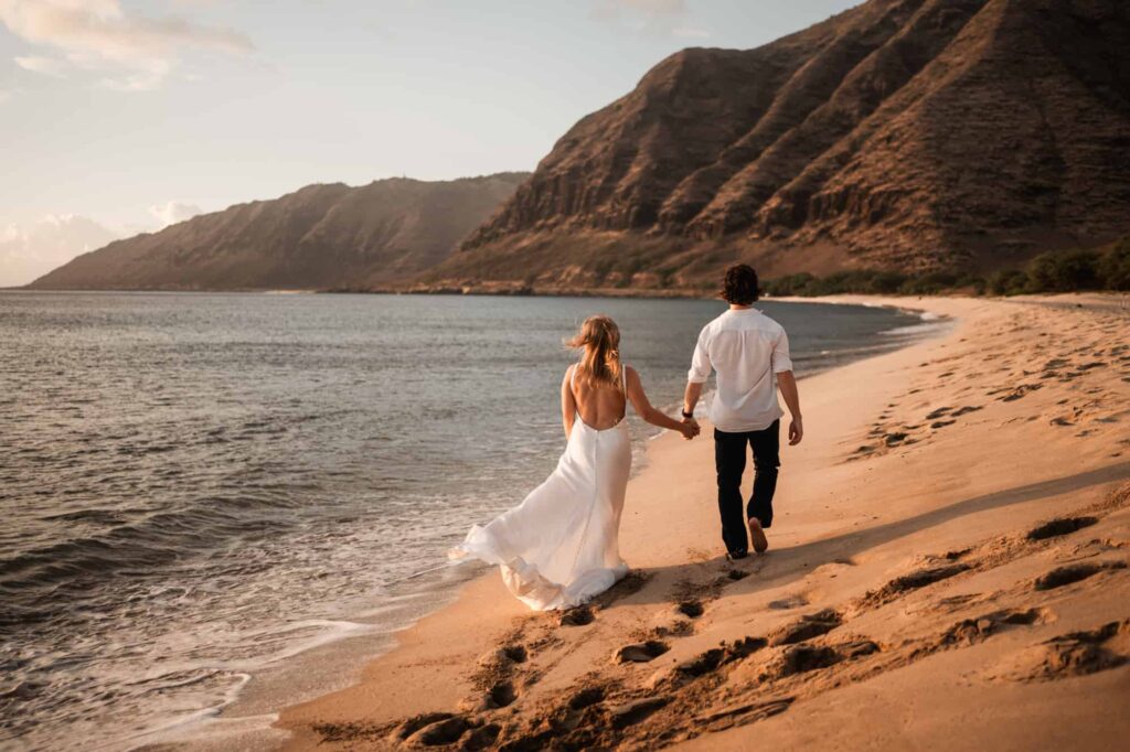 A destination wedding photographer captures a serene moment of a couple walking hand-in-hand along a deserted Hawaiian beach at sunset, with the bride in an elegant white dress and the groom in a casual white shirt and black pants, against a backdrop of gentle waves and rolling hills.