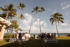 Outdoor destination wedding ceremony in Hawaii, with guests gathered on a lush green lawn facing the ocean. Tall palm trees sway above, framing a clear blue sky. White chairs line the aisle leading to the bride and groom at the altar near the sea