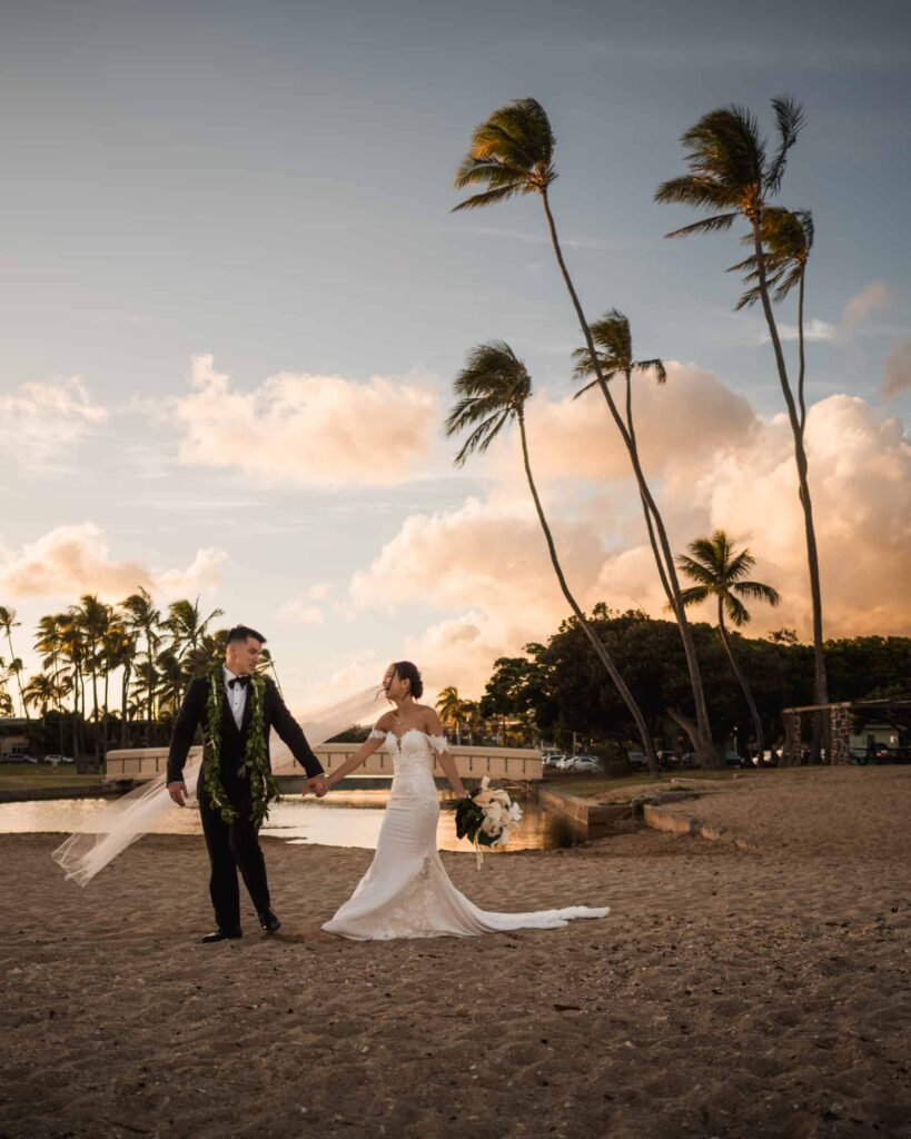 A couple at their destination wedding in Hawaii, with the bride in a chic form-fitting dress and long veil and the groom in a classic black tuxedo, holding hands and walking on a sandy beach with tall palm trees swaying in the breeze against a sunset sky.