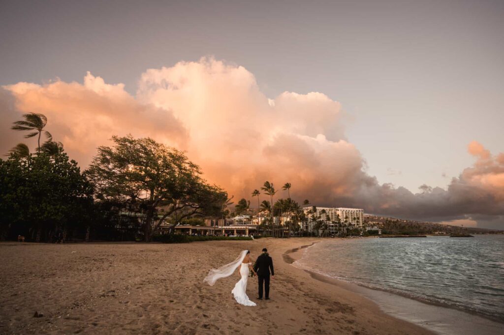 A breathtaking Hawaii beach at dusk sets the scene for a destination wedding, where the bride in a long white gown with a trailing veil and the groom in a dark suit stand hand in hand, looking at the dramatic pink and orange clouds over the calm sea, with palm trees framing the picturesque resort in the background