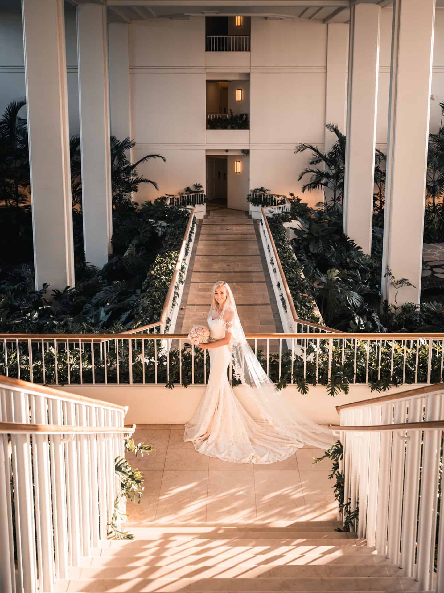 Bride in a detailed lace wedding dress and flowing veil holding a bouquet, posing on a grand staircase with lush greenery and white architectural columns at a luxurious Hawaiian resort, as the warm sunset light enhances the romantic atmosphere