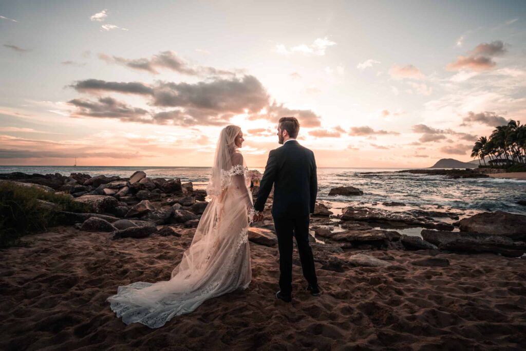 An elopement photographer in Oahu, Hawaii, captures an intimate sunset scene with a couple standing on a rocky beach, holding hands and gazing into each other’s eyes, with the bride’s lace-trimmed veil trailing in the sand and the groom in a sharp black suit, set against a stunning backdrop of the ocean and a pastel sky.