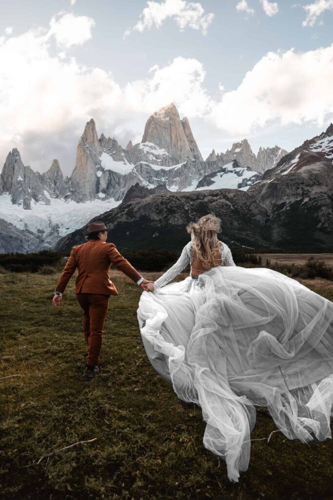 Destination wedding photographer captures a couple holding hands in a vast meadow with the majestic, snow-capped peaks of Mount Fitz Roy in the background, showcasing the bride's flowing gown and the groom's stylish brown suit.