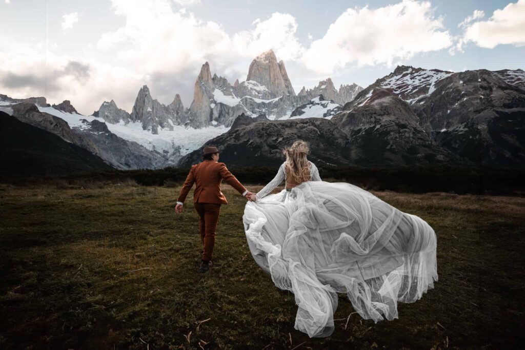 Destination wedding photographer captures a couple holding hands in a vast meadow with the majestic, snow-capped peaks of Mount Fitz Roy in the background, showcasing the bride's flowing gown and the groom's stylish brown suit