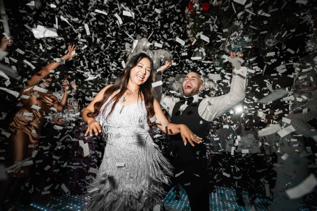 Joyful moment captured by a destination wedding photographer as a couple dances under a shower of confetti, with the bride in a shimmering pink fringe dress and the groom in a classic black tuxedo, both laughing heartily amidst a festive atmosphere