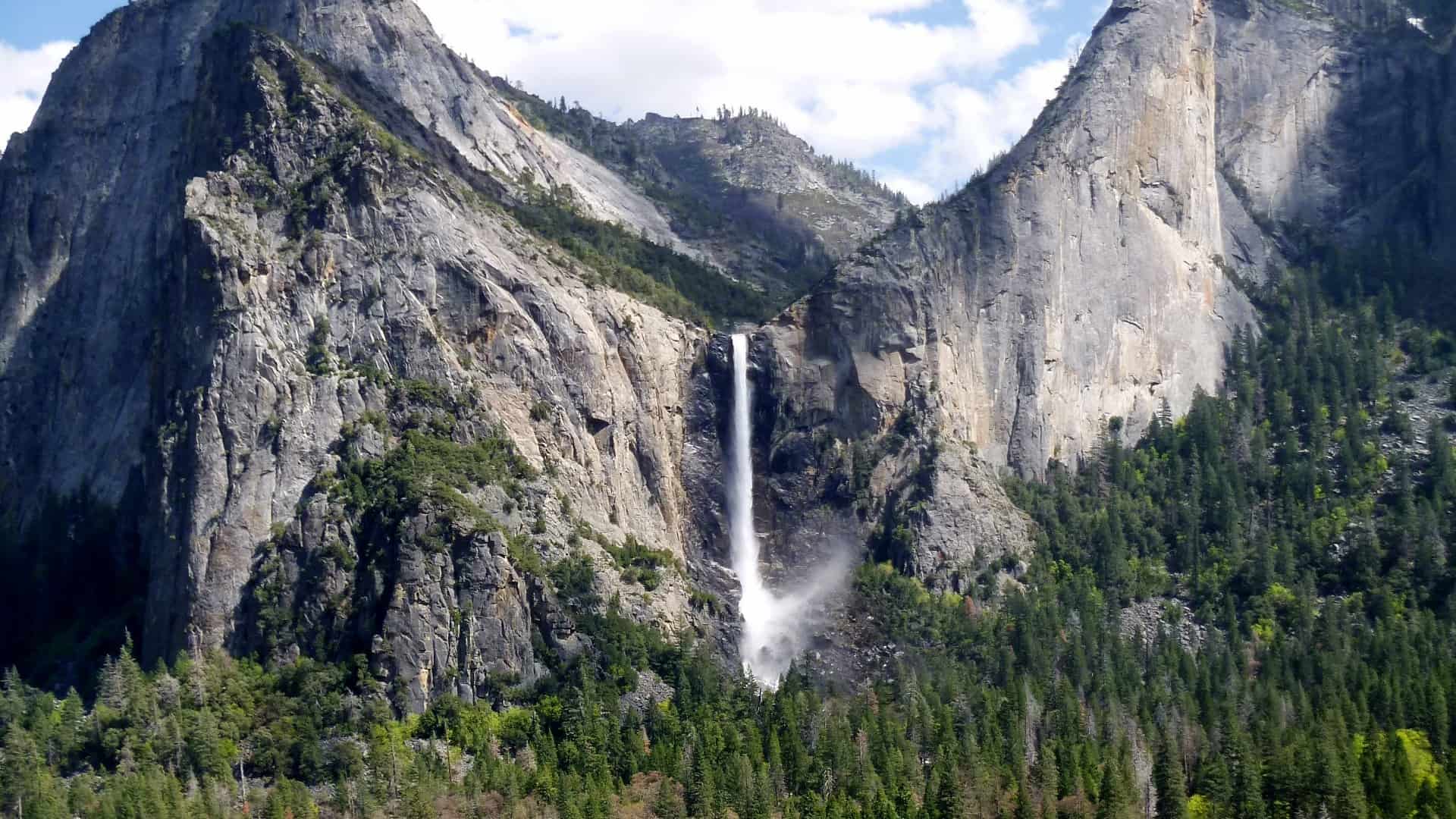 Bridalveil Fall in Yosemite National Park, a stunning waterfall plunging from the granite cliffs surrounded by pine forests, with mist rising from the base of the fall, set against a background of rugged mountain terrain