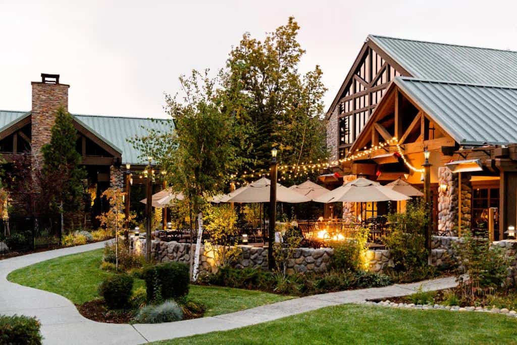 Twilight at Tenaya Lodge near Yosemite with string lights illuminating the outdoor patio area, featuring stone fire pits, cozy seating under umbrellas, amidst landscaped gardens with a meandering path, inviting guests to a warm and rustic mountain resort experience