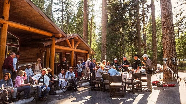 Guests enjoy a sunny outdoor gathering at The Redwoods in Yosemite, with a rustic wooden lodge in the background, set among tall pine trees, creating a cozy and inviting atmosphere for socializing in the heart of the national park