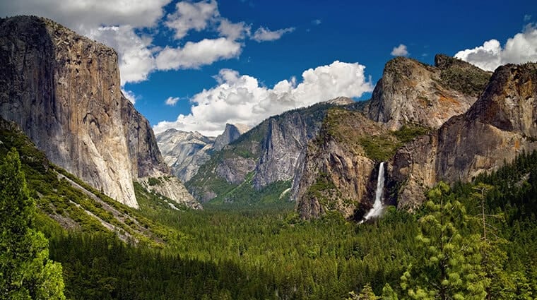 The iconic Tunnel View in Yosemite, offering a breathtaking panorama of the valley with El Capitan on the left, Bridalveil Fall on the right, and the Half Dome in the distant center, under a sky dotted with fluffy clouds
