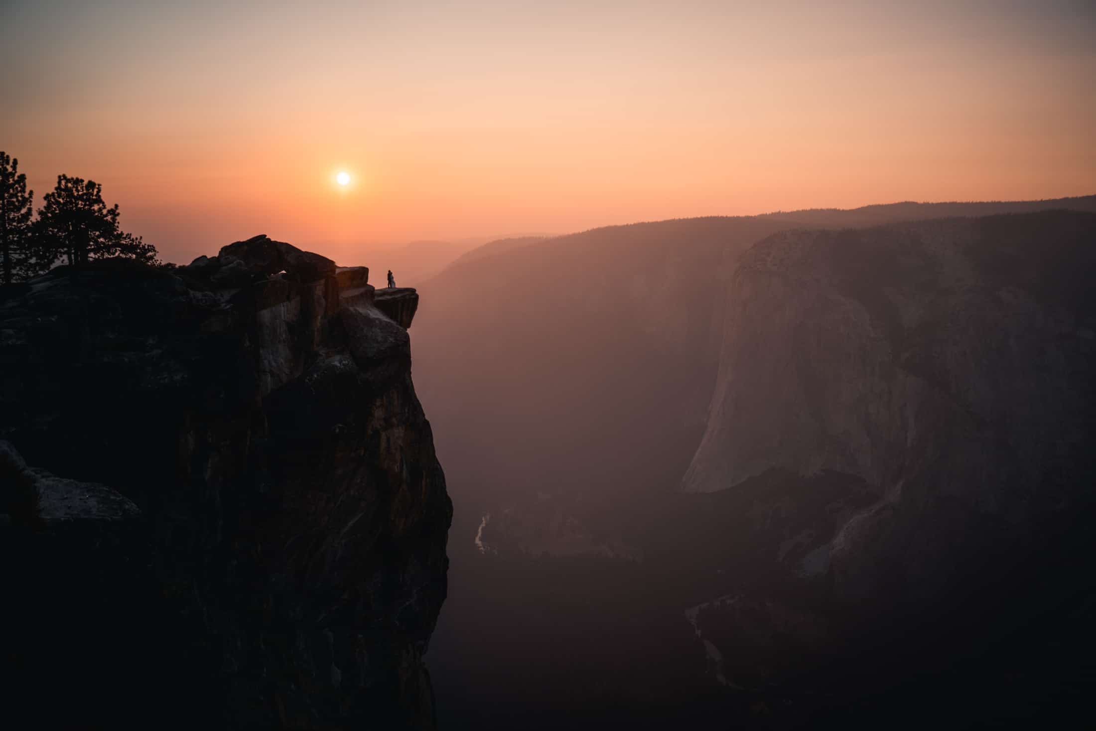A solitary figure stands on the edge of Taft Point in Yosemite at sunset, overlooking the vast valley with the sun casting a warm glow over the granite cliffs and the silhouette of the distant mountains