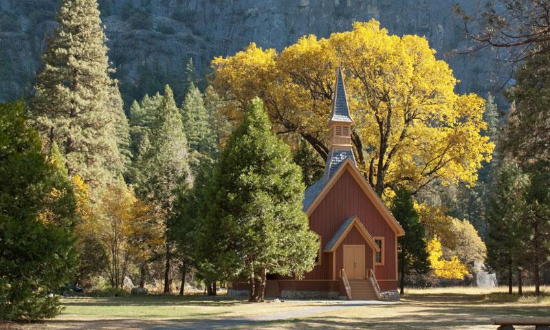 Alt text: "The Yosemite Valley Chapel in autumn, a quaint brown wooden church with a tall steeple, set against a backdrop of towering evergreens and a striking golden-leafed tree, encapsulating the serene beauty of the historic site within Yosemite National Park