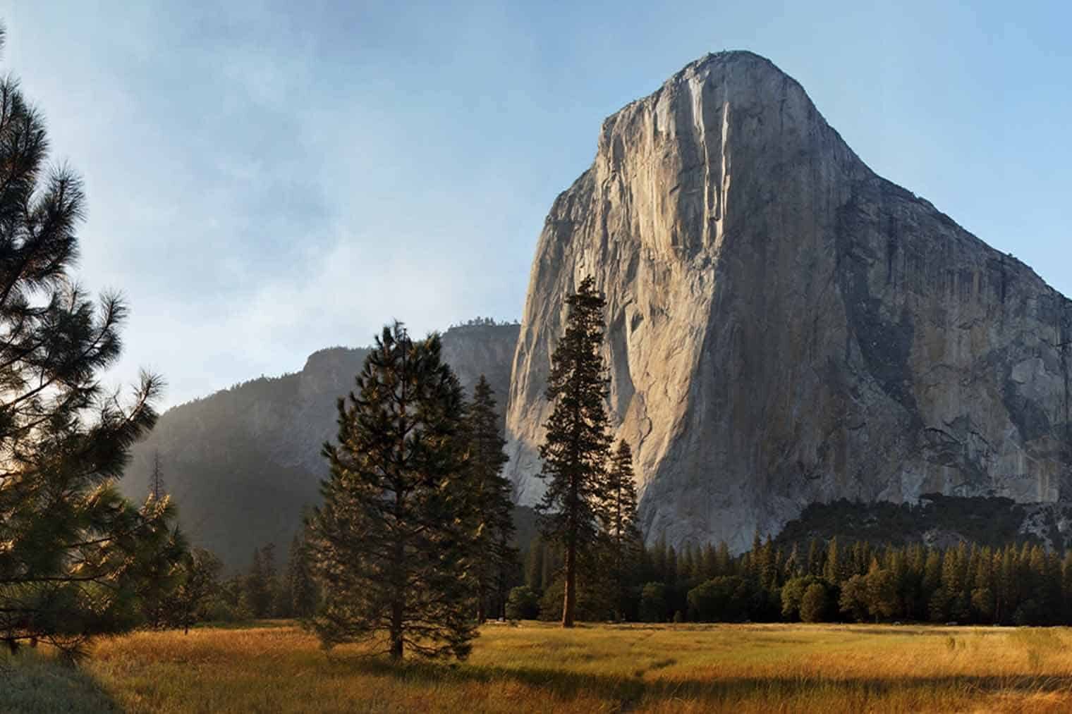 The majestic El Capitan Meadow in Yosemite, with the iconic sheer face of El Capitan rising in the background, a clear blue sky above, and a peaceful meadow with a footbridge and visitors enjoying the idyllic outdoor setting