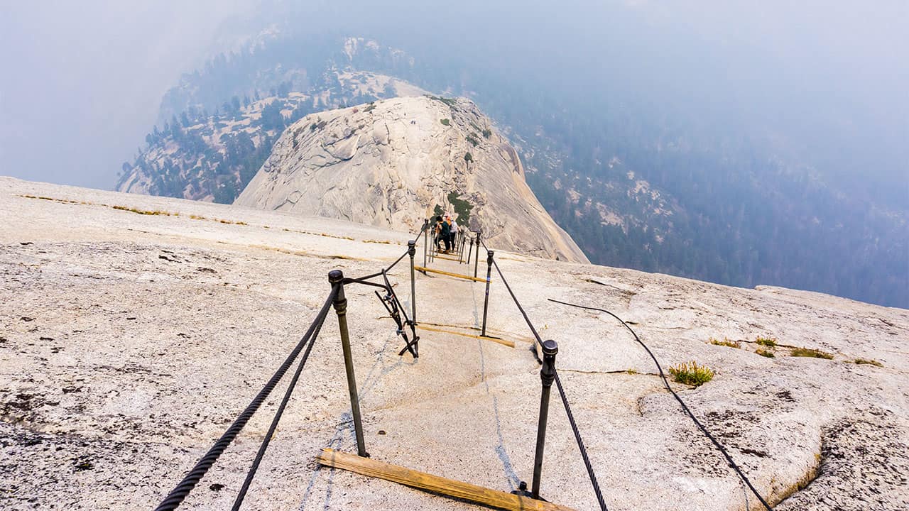 Hikers navigate the steep granite ascent of Half Dome in Yosemite using the secured cables, showcasing the challenging and rewarding climb against a backdrop of hazy skies and forested valley views