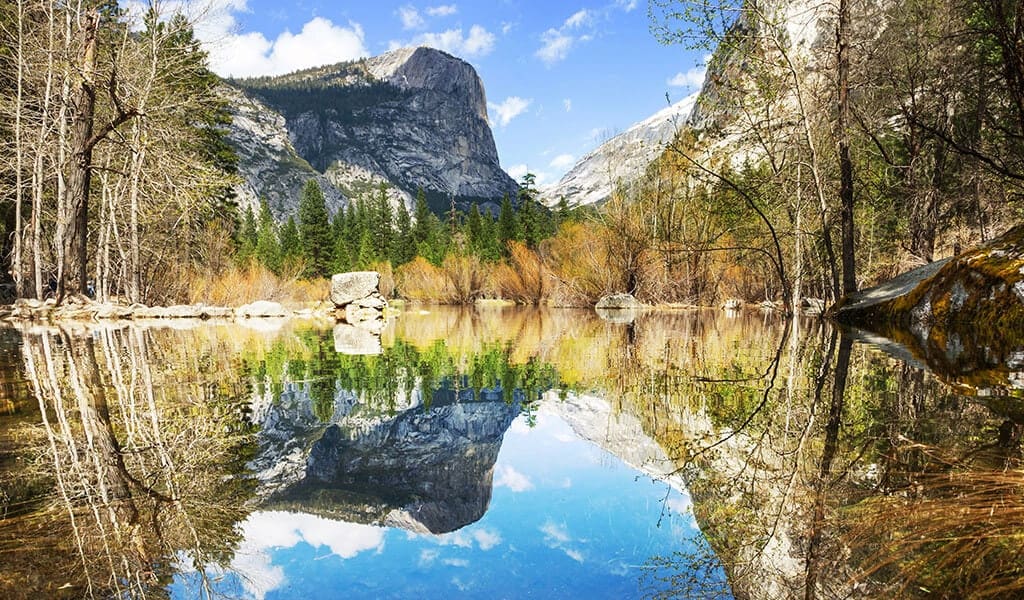 Mirror Lake in Yosemite National Park reflects the clear blue sky and the imposing granite cliffs, including a perfect reflection of Half Dome, surrounded by serene waters and forested banks.