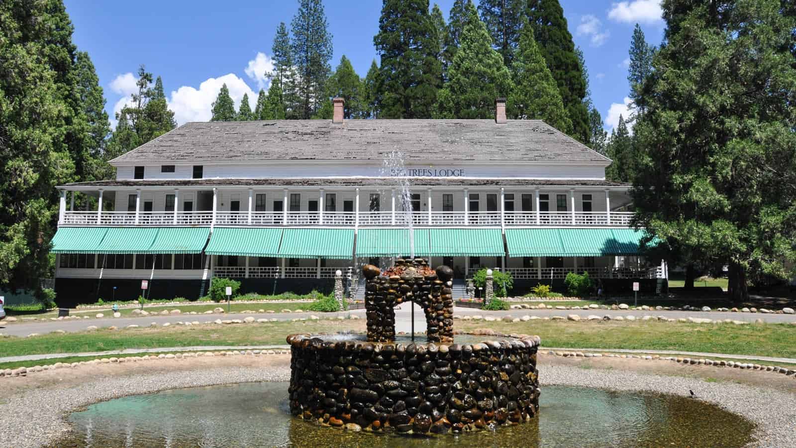 Historic Wawona Hotel in Yosemite National Park, a charming two-story structure with white walls and green trim, featuring a stone fountain in the foreground, surrounded by lush greenery and towering trees under a clear blue sky
